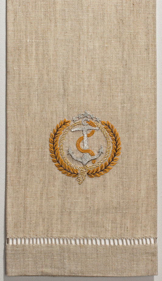 A flax colored hand towel with a hemstitch. Embroidered on it is a silver anchor with a gold laurel wreath circling it.