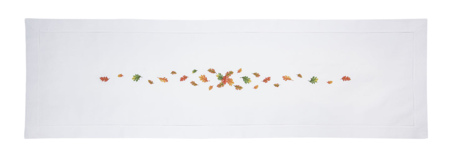 A white runner with a hemstitch border. Embroidered in a line down the center is bunch of multicolored fall leaves