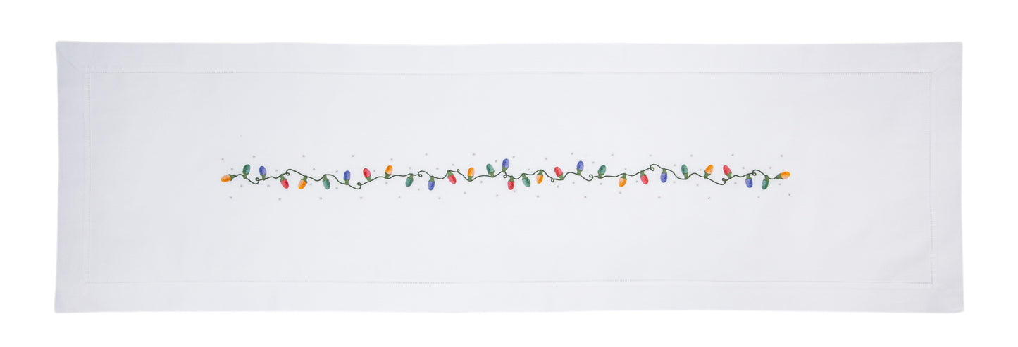 A white table runner with a hemstitch border. A string of holiday lights in red yellow green & blue are in a line down the middle.