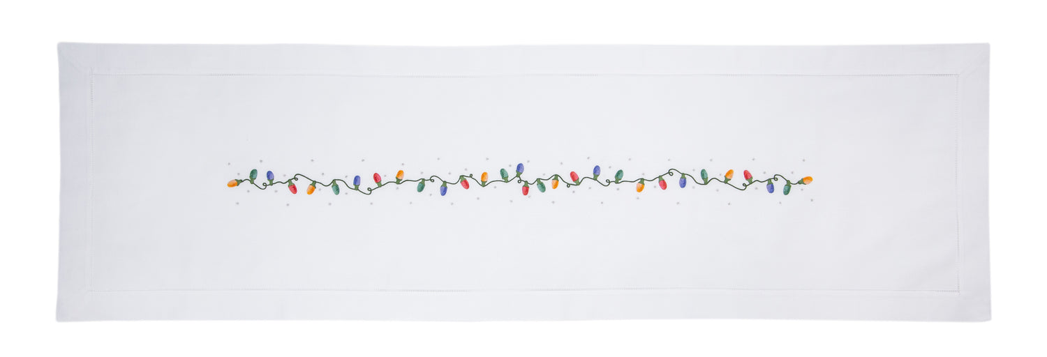 A white table runner with a hemstitch border. A string of holiday lights in red yellow green & blue are in a line down the middle.