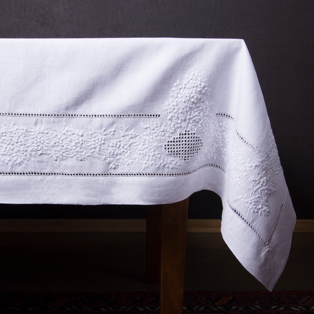 A white linen tablecloth with a hemstitch border and white french knot floral embroidery draped off a table