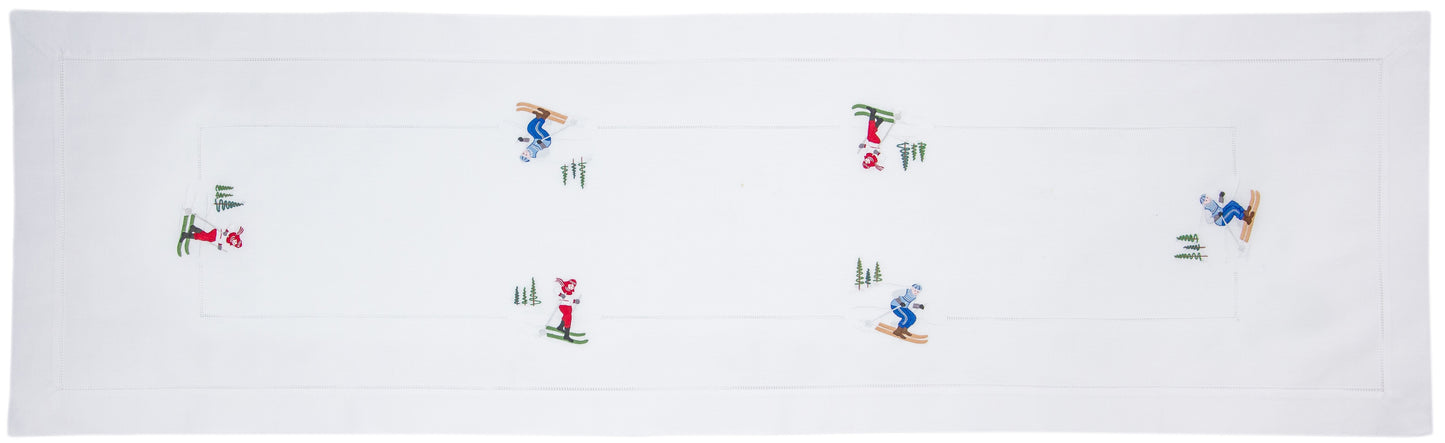 A white table runner with a hemstitch border. Skiers are embroidered in a border around the edge