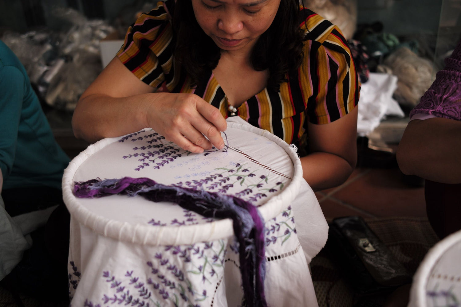 Woman in striped shirt sitting on the ground hand-embroidering white cloth with purple design