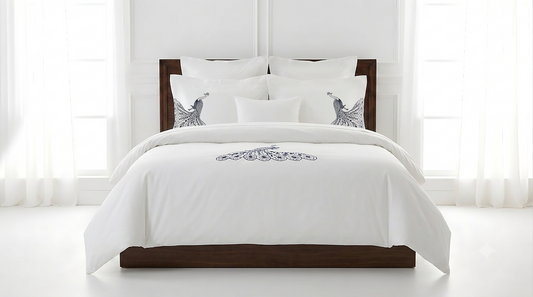 A white comforter set on a dark wood bed frame, styled with multiple white pillows, including two decorative pillows featuring a blue and gray bird design, set against a bright white window background.