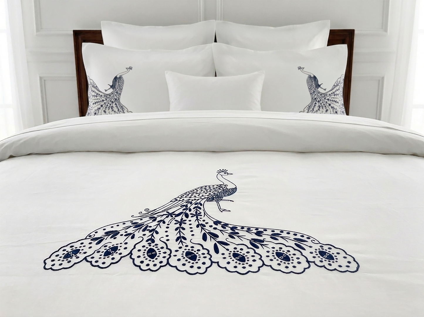 A neatly made bed with white bedding featuring a large navy blue peacock illustration on the duvet and matching peacock motifs on two pillowcases, set against a white paneled headboard.