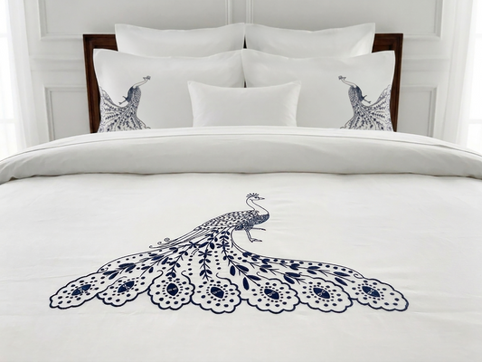 A neatly made bed with white bedding featuring a large navy blue peacock illustration on the duvet and matching peacock motifs on two pillowcases, set against a white paneled headboard.
