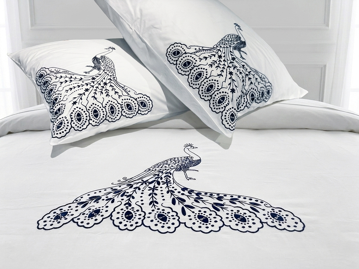 White bedding set with a large navy blue peacock print on the duvet cover and matching pillowcases, styled on a bed against a white headboard.