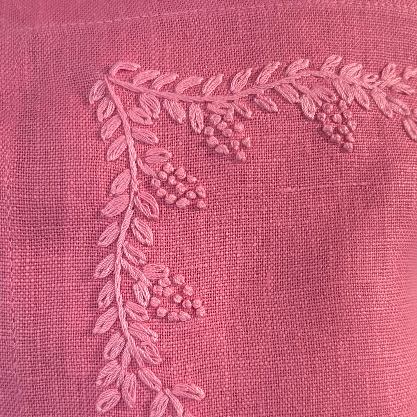 Close-up of warm pink fabric with a raised embroidered leafy branch and small berry clusters in a lighter pink thread.