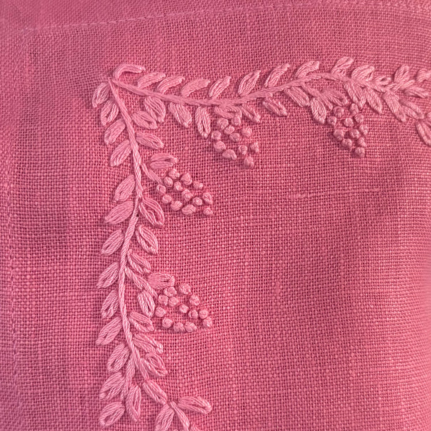 Close-up of warm pink fabric with a raised embroidered leafy branch and small berry clusters in a lighter pink thread.
