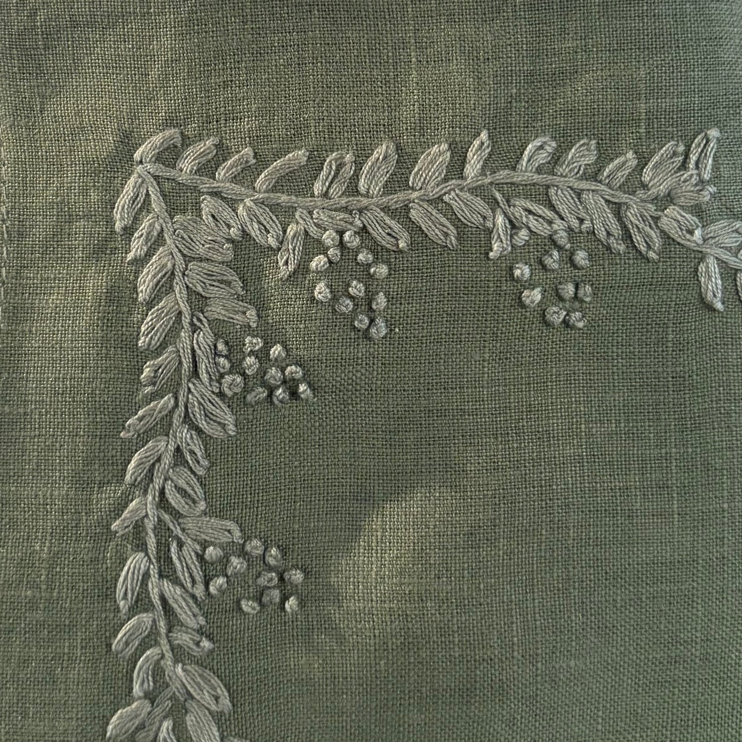 Close-up of olive green fabric with a raised embroidered leafy branch and small berry clusters in a lighter gray thread.