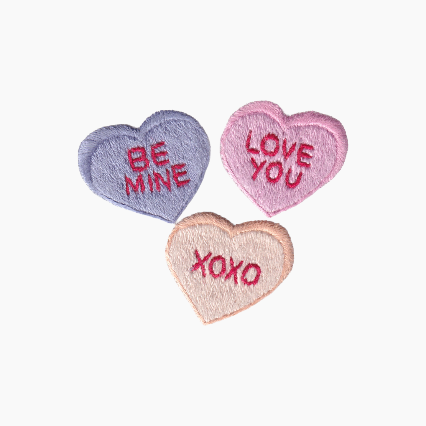 Hand embroidered cotton boutique tissue box cover of three heart-shaped candy with embroidered text 'Be Mine', 'Love You', and 'XOXO' on a white background.