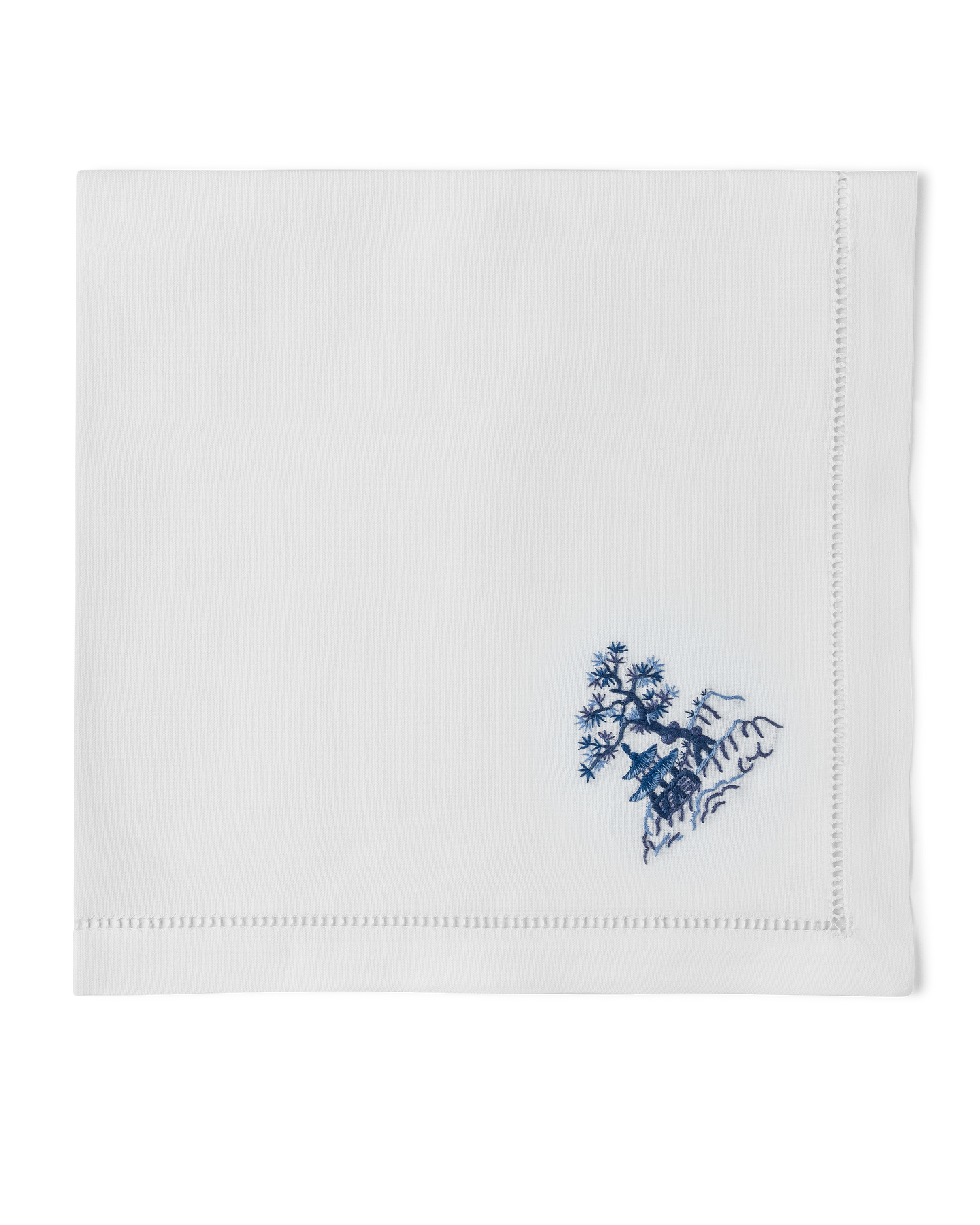 A white napkin with a hemstitch. A house & tree on a hill are embroidered in blue in the bottom righthand corner.