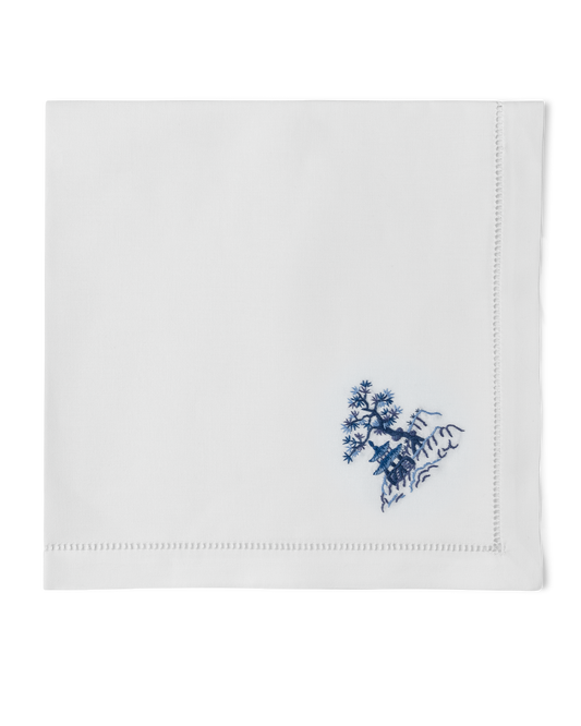 A white napkin with a hemstitch. A house & tree on a hill are embroidered in blue in the bottom righthand corner.
