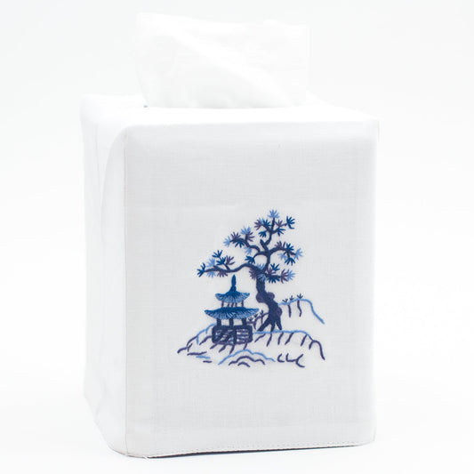 A white tissue box cover with a house & tree on a hill embroidered in blue in the center.