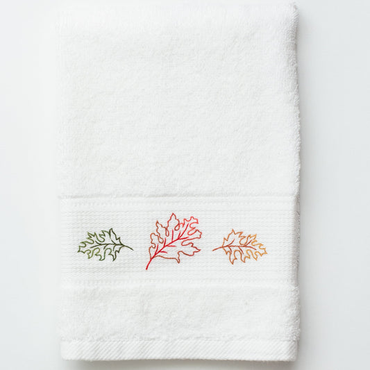 A white terry towel with outlines of 3 of fall leaves in green, orange and red embroidered in the center.