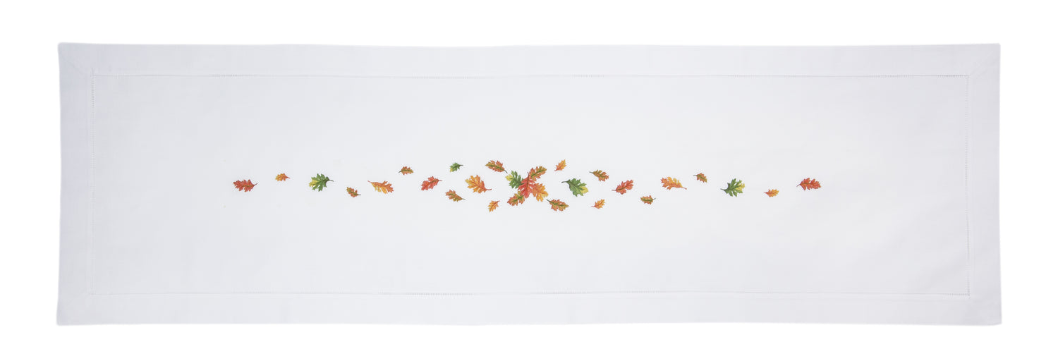 A white runner with a hemstitch border. Embroidered in a line down the center is bunch of multicolored fall leaves