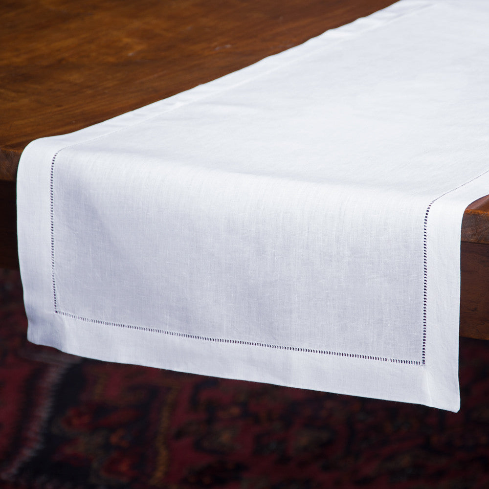 A white linen table runner with a hemstitch border, draped off the end of a wooden table.