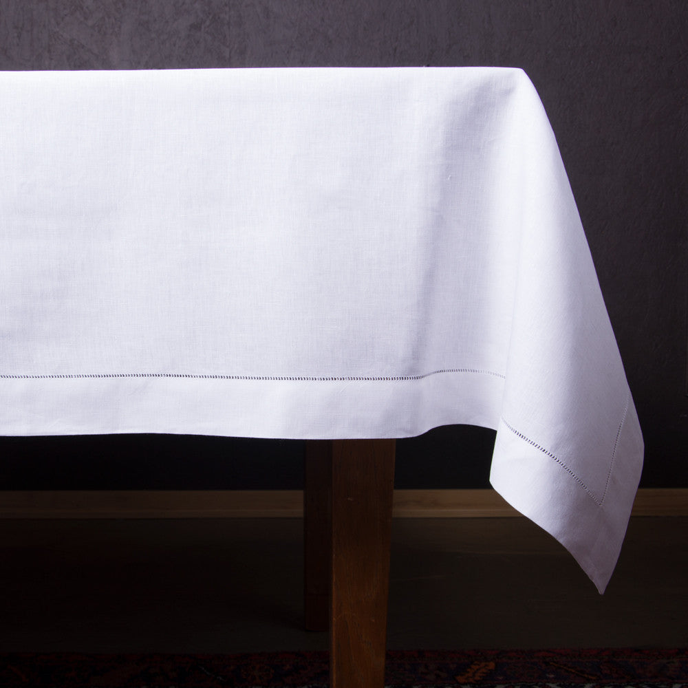 A white linen tablecloth with a hemstitch border draped over the side of a table (side view)