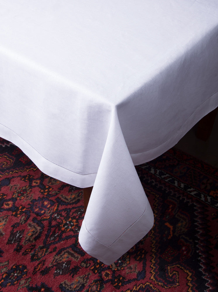 A white linen tablecloth with a hemstitch border draped over the side of a table (corner view)