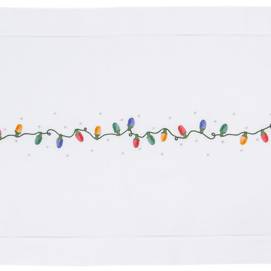 A white table runner with a hemstitch border. A string of holiday lights in red yellow green & blue are in a line down the middle.