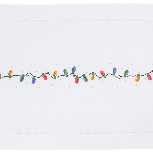 A white table runner with a hemstitch border. A string of holiday lights in red yellow green & blue are in a line down the middle.