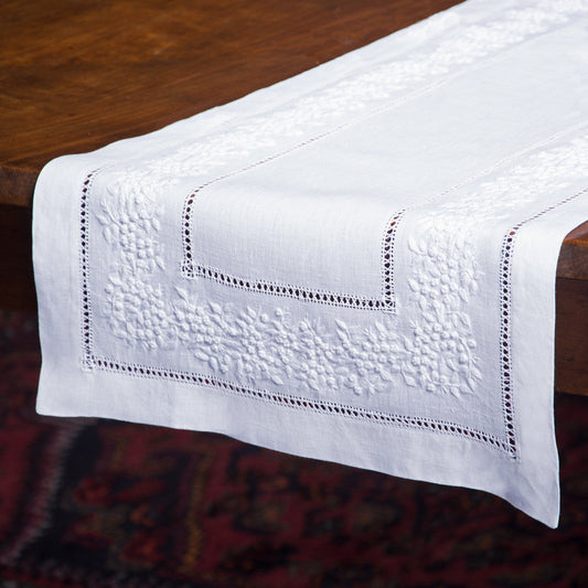 A close up of a white linen table runner with a hemstitch border draped off the edge of a wooden table