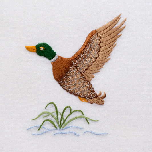 A white hand towel with a hemstitch. A mallard flying above water is embroidered in the center