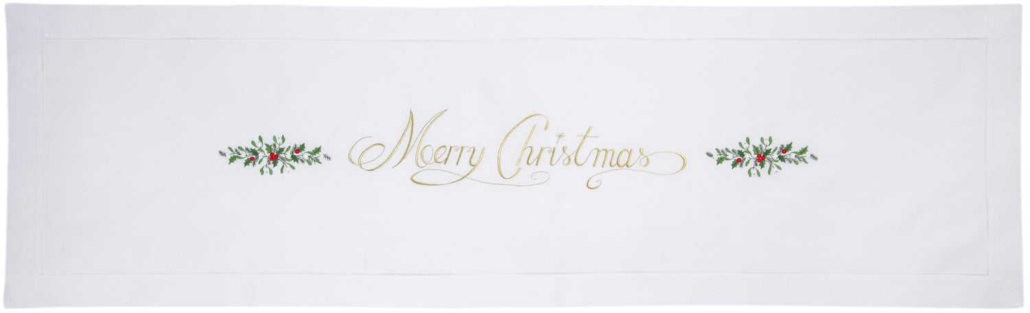 A white table runner with a hemstitch border. The words “Merry Christmas” in gold with holly accents runs down the middle