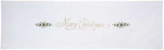 A white table runner with a hemstitch border. The words “Merry Christmas” in gold with holly accents runs down the middle