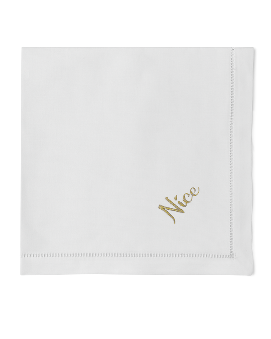 A white napkin with a hemstitch. The word “nice” in gold is embroidered in the bottom right corner