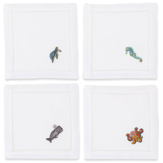 4 white cocktail napkins with a hemstitch border. Embroidered on the bottom right corner of each is a whale, seahorse, octopus, and turtle