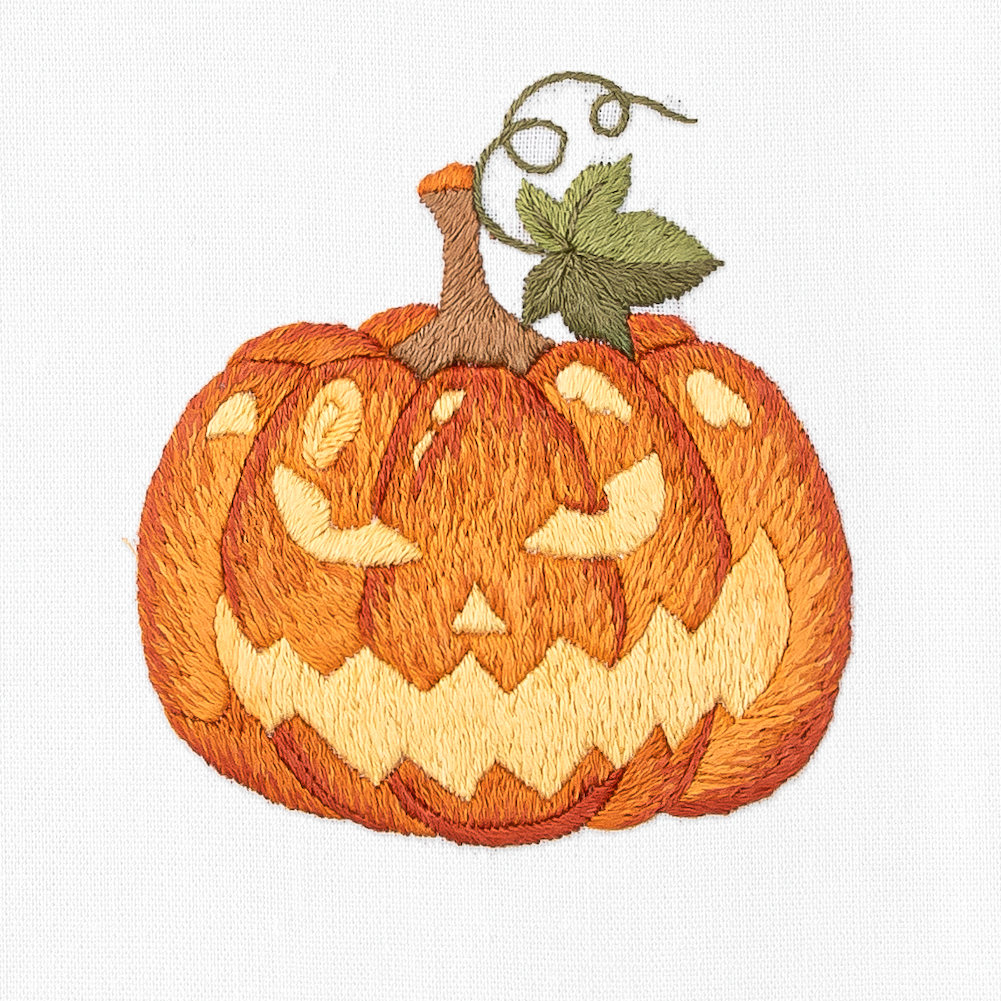 A close up detailed image of the embroidery - A jack o lantern with yellow detailing and a green leaf next to the stem