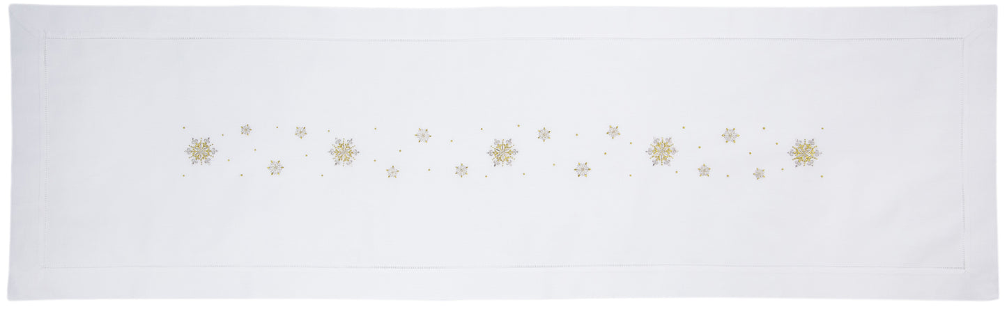 A white table runner with a hemstitch border. Gold & silver metallic snowflakes are embroidered in a line down the middle