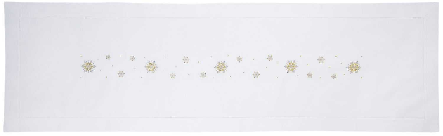 A white table runner with a hemstitch border. Gold & silver metallic snowflakes are embroidered in a line down the middle